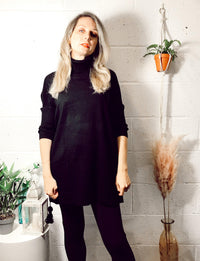 Woman wearing a black Edie roll-neck tassel trim poncho with long sleeves, styled with black leggings in a cozy indoor setting featuring potted plants and decorative accents.