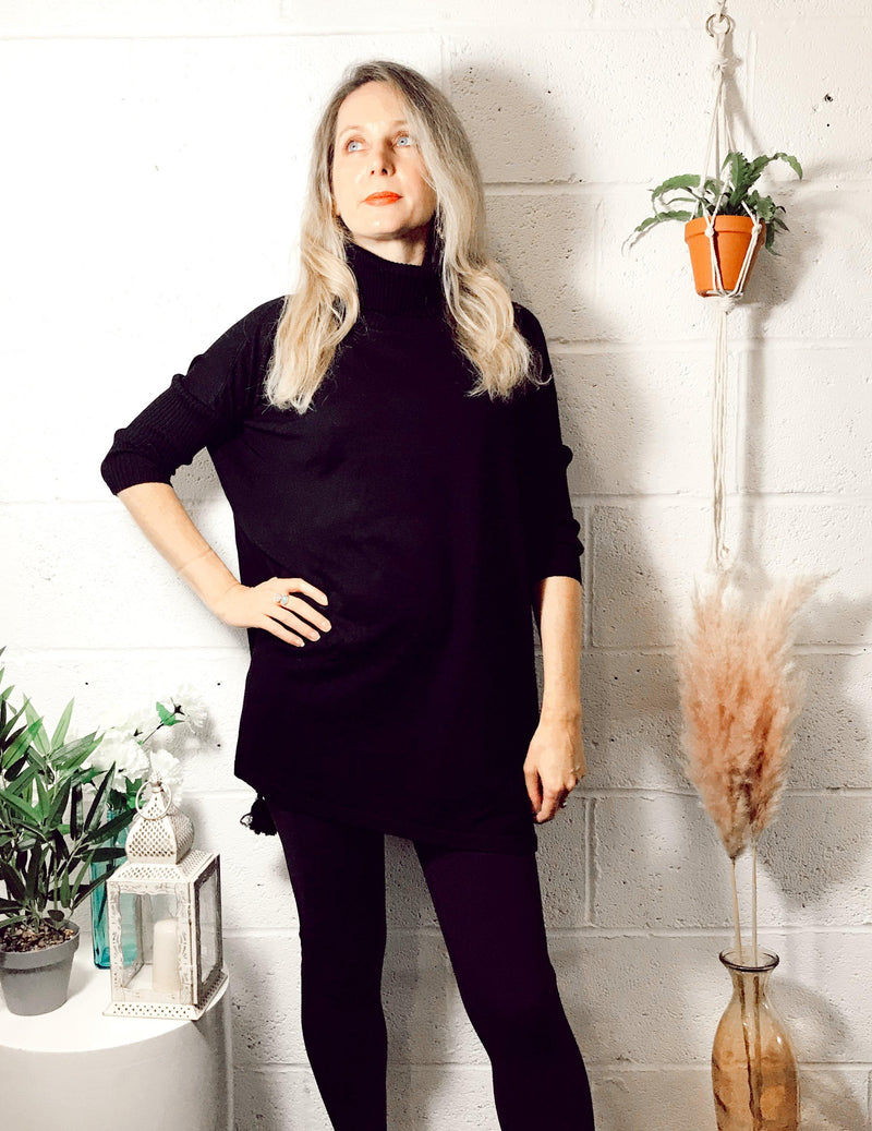 Woman wearing a black Edie roll-neck tassel trim poncho with three-quarter sleeves and a high neckline, standing confidently against a white textured wall with plants nearby.