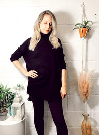 Woman with blonde hair wearing a black Edie roll-neck tassel trim poncho, paired with black leggings, standing indoors next to plants and decorative items.