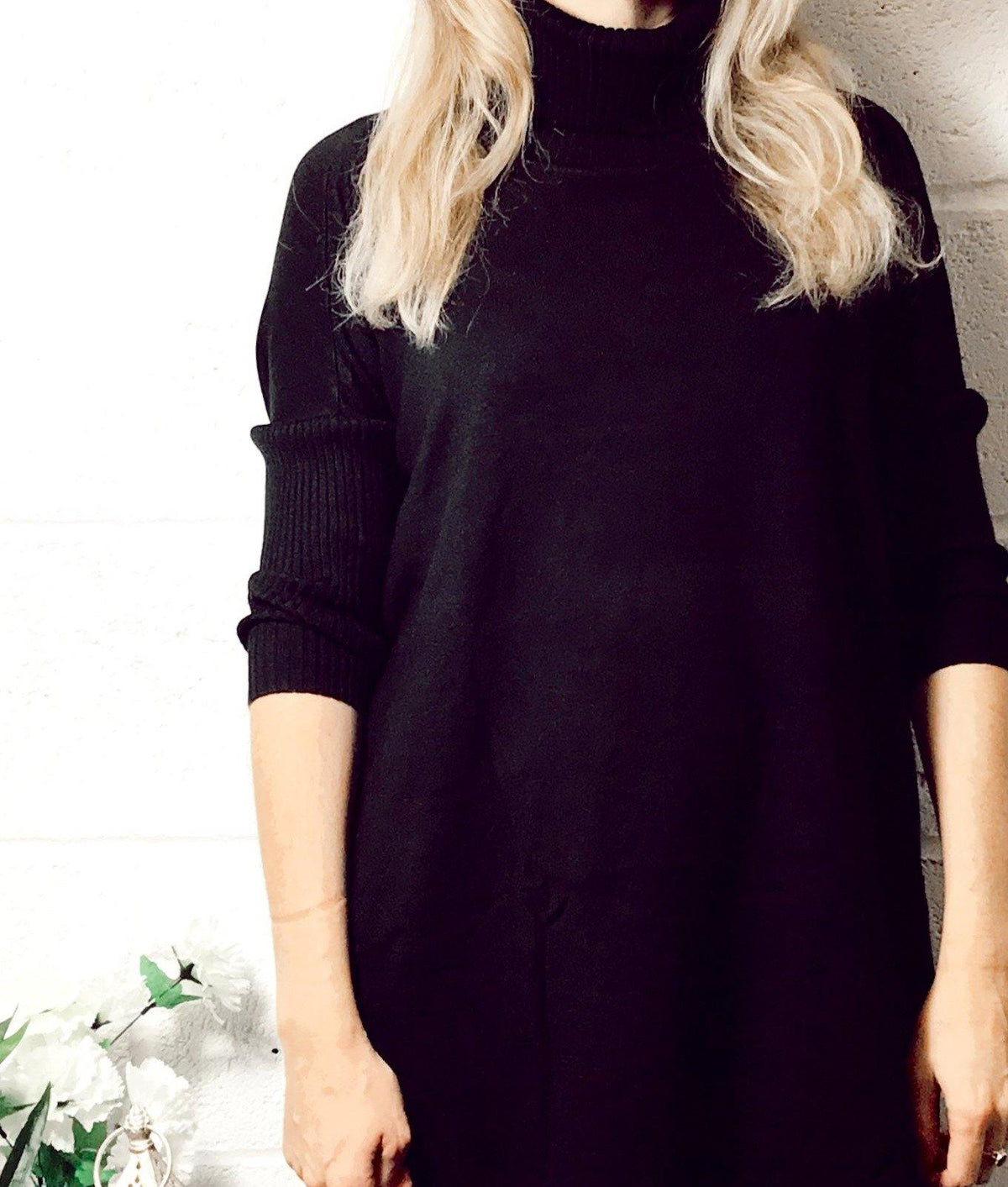 Woman wearing a black Edie roll-neck tassel trim poncho with long sleeves and a cozy turtleneck in a minimalist setting.