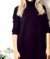 Woman wearing a black Edie roll-neck tassel trim poncho with long sleeves and a cozy turtleneck in a minimalist setting.