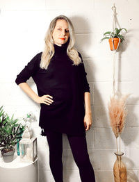 Woman wearing a black Edie roll-neck tassel trim poncho standing indoors by a white wall with plants and decor, showcasing a comfortable boho style outfit.