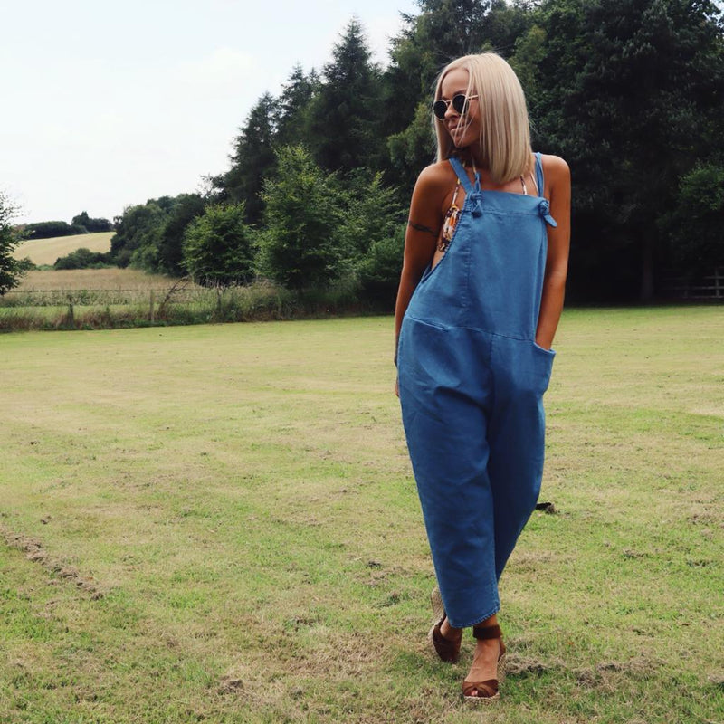 Woman wearing a Denim light-blue jumpsuit//dungarees standing on green grass with trees in the background, paired with sunglasses and sandals in a relaxed outdoor setting.