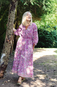 Woman smiling outdoors wearing a Rosalie bohemian floral-print pink cotton maxi kaftan//dress free size with long sleeves and relaxed fit, standing by a tree in a sunlit forest path.