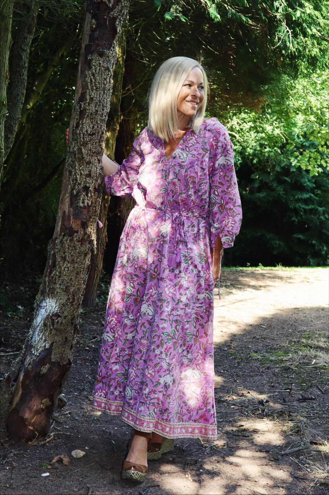 Woman wearing a Rosalie bohemian floral-print pink cotton maxi kaftan//dress free size, standing outdoors near a tree in a sunlit natural setting with greenery in the background.