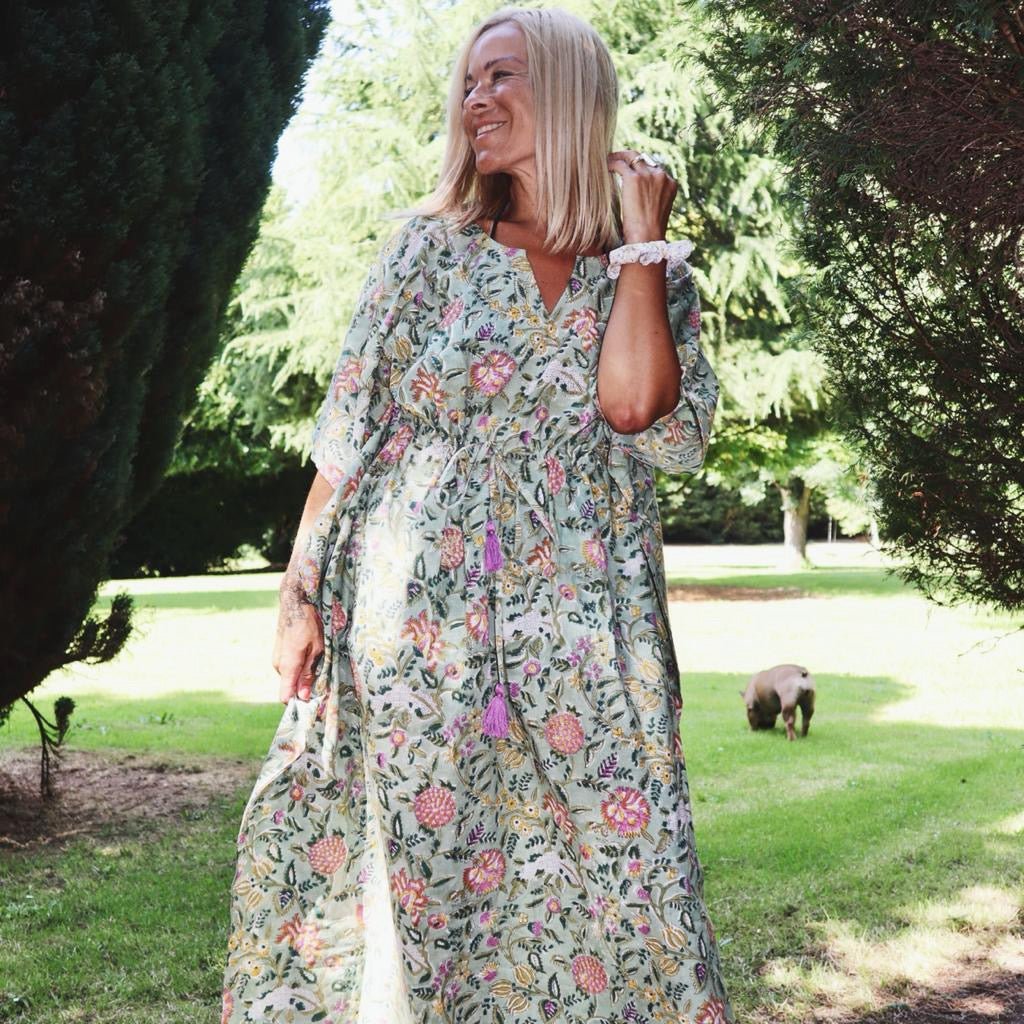 Woman wearing Eden bohemian floral-print green cotton maxi kaftan//dress free size standing outdoors in a sunlit garden surrounded by trees, smiling.