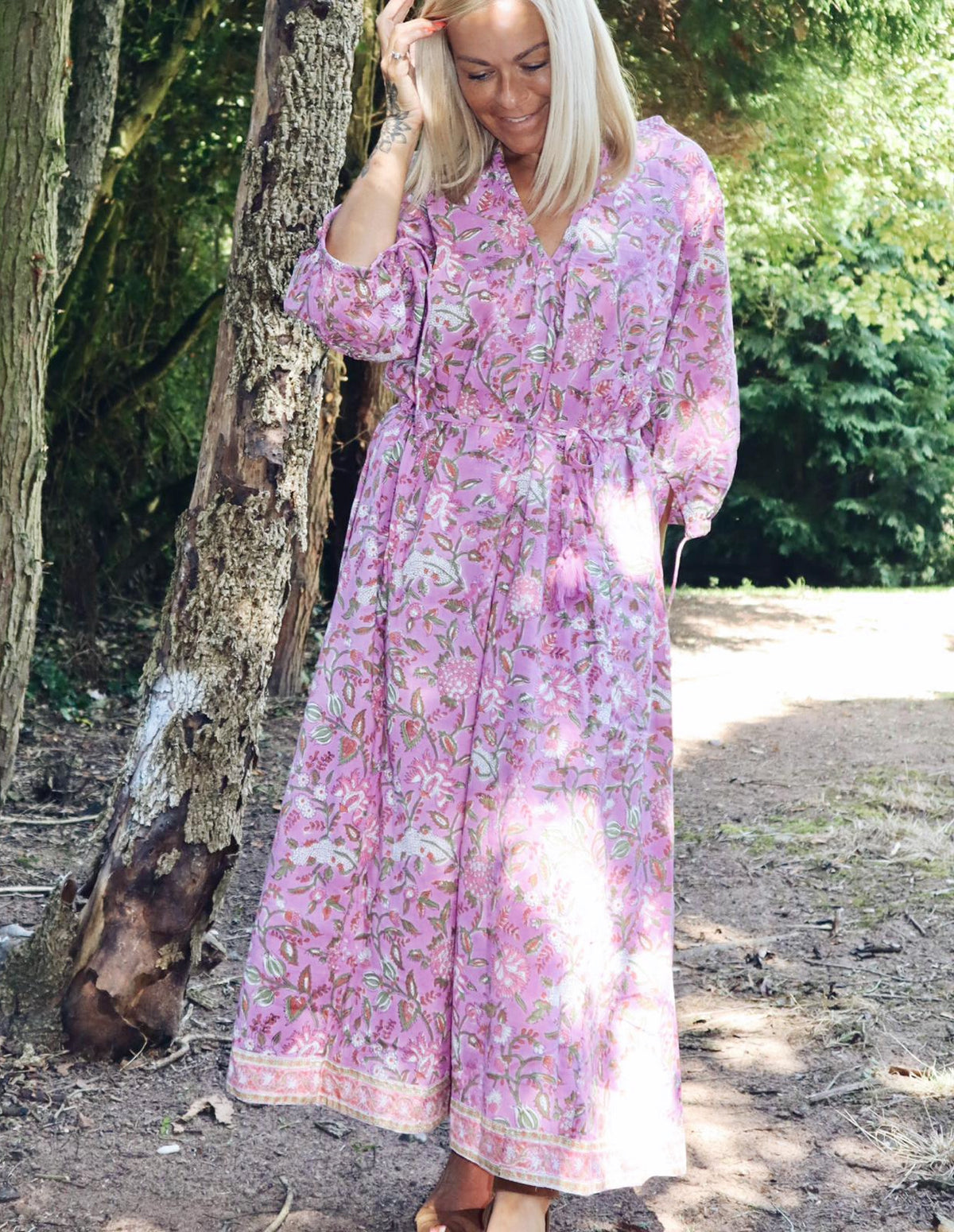 Woman wearing Rosalie bohemian floral-print pink cotton maxi kaftan//dress free size standing outdoors near a tree in a sunlit natural setting.