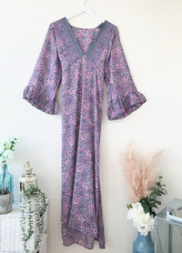 Bluebell lilac vintage-silk free-size ruffled-sleeved tie maxi dress hanging on a white hanger with a paisley boho pattern in soft lilac and blue tones against a neutral background with decorative plants and lantern nearby.