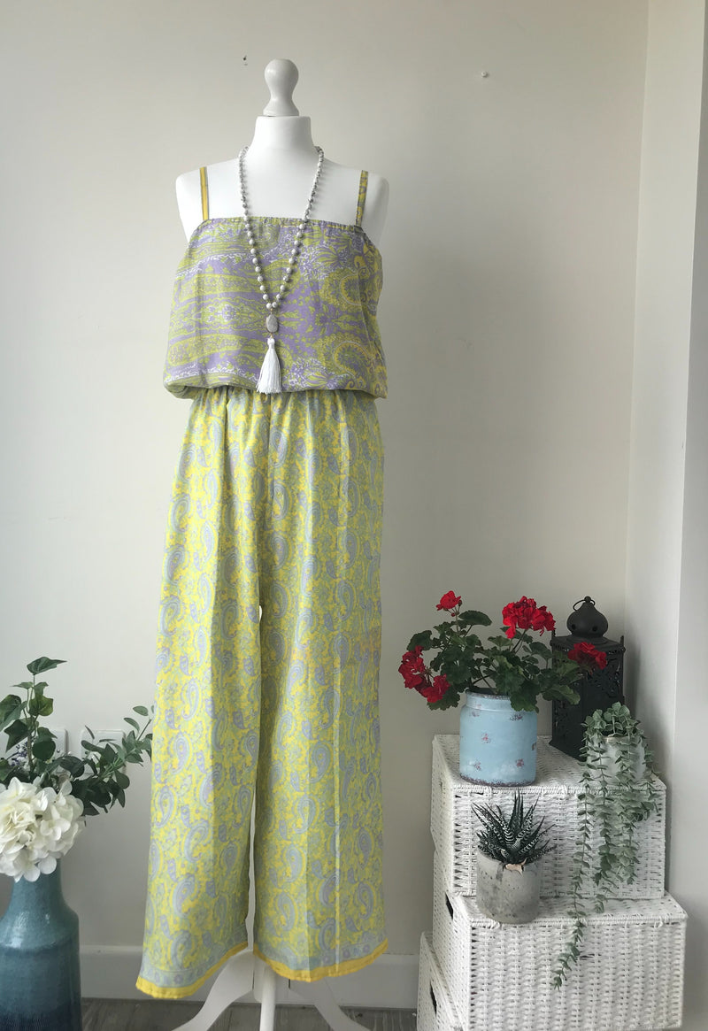 Mannequin wearing a Layla yellow/lilac paisley-print silk jumpsuit free-size with a layered tassel necklace, displayed next to white wicker furniture and potted plants in a softly lit room.