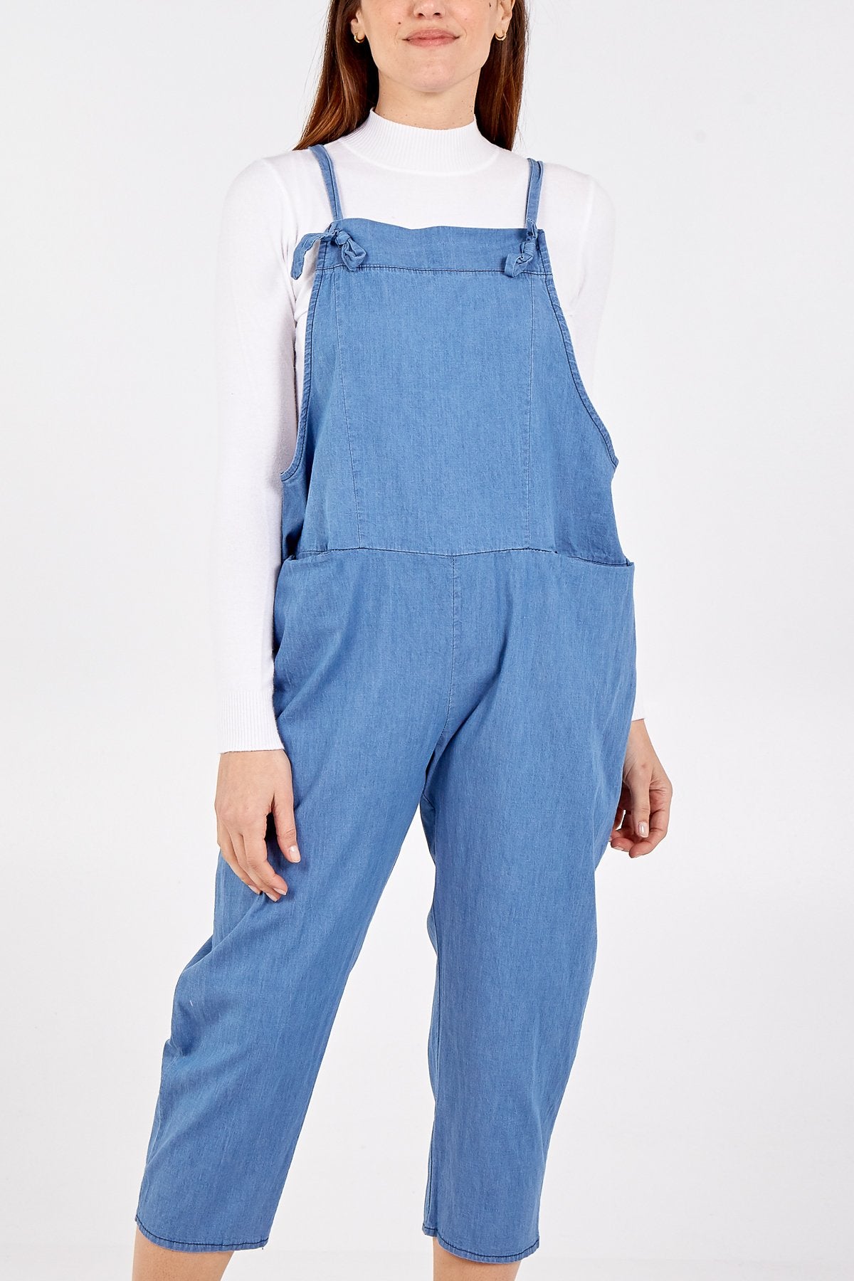 Woman wearing a Denim light-blue jumpsuit//dungarees over a white long-sleeve top, featuring adjustable shoulder straps and a relaxed fit.