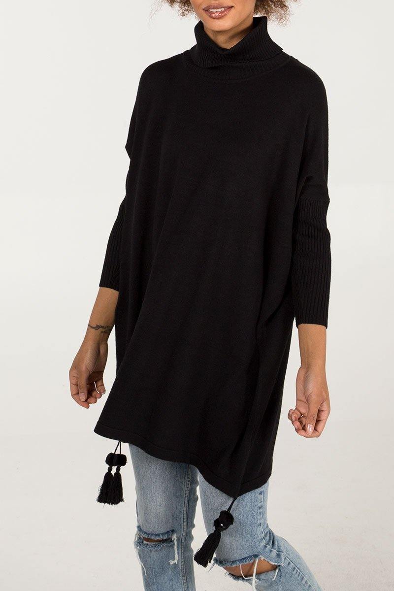Woman wearing a black Edie roll-neck tassel trim poncho with long ribbed sleeves and tassel details, styled with ripped light blue jeans in a casual boho look.