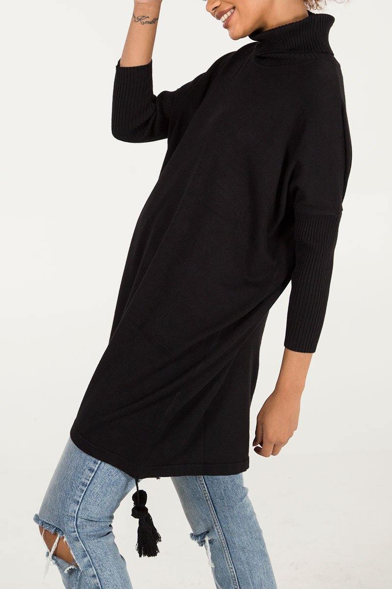 Woman wearing a black Edie roll-neck tassel trim poncho over distressed light blue jeans, featuring a high turtleneck and relaxed fit with tassel detail at the hem.