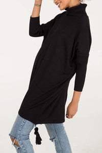 Woman wearing a black Edie roll-neck tassel trim poncho over distressed light blue jeans, featuring a high turtleneck and relaxed fit with tassel detail at the hem.