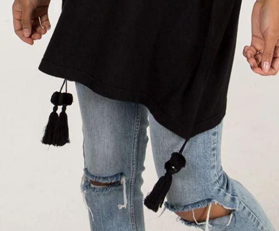 Close-up of a person wearing light blue ripped jeans paired with a black Edie roll-neck tassel trim poncho featuring tassels hanging at the hem.